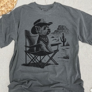 Pu&ograve; includere: T-shirt grigio con grafica nera di un cane con cappello da cowboy e bandana, seduto su una sedia pieghevole. Il design include un paesaggio desertico con un cactus e mesas. La maglietta &egrave; in materiale morbido.