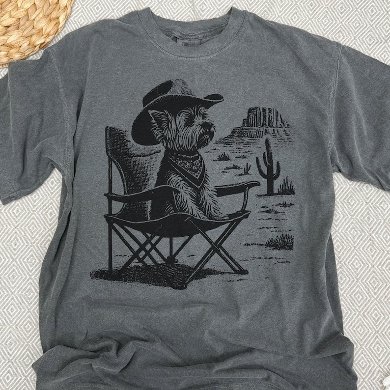 Pu&ograve; includere: T-shirt grigio con grafica nera di un cane con cappello da cowboy e bandana, seduto su una sedia pieghevole. Il design include un paesaggio desertico con un cactus e mesas. La maglietta &egrave; in materiale morbido.