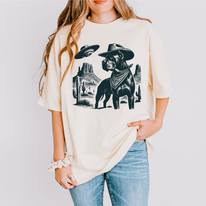 May include: Cream-colored t-shirt featuring a black and white graphic of a dog wearing a cowboy hat and bandana, with a UFO and desert landscape. The shirt has a relaxed fit.