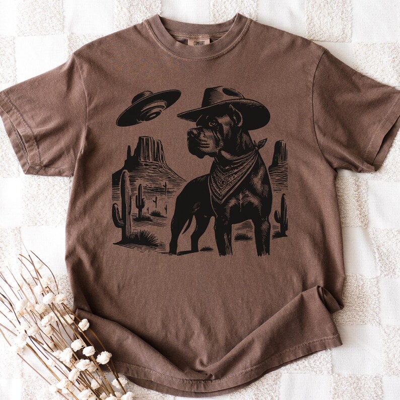 May include: Brown t-shirt featuring a black graphic of a dog wearing a cowboy hat and bandana, with a UFO and desert landscape. The shirt has a crew neck and short sleeves.
