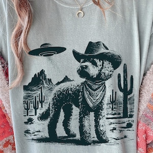 Peut inclure: T-shirt bleu clair avec un motif noir et blanc d'un chien portant un chapeau de cowboy et un bandana, avec un OVNI au-dessus d'un paysage désertique avec des cactus et des montagnes.