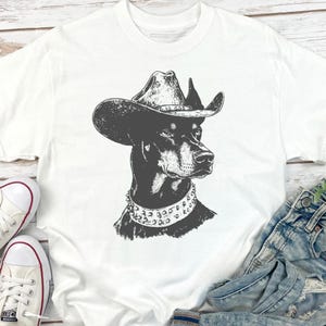 Pu&ograve; includere: T-shirt bianco con un'illustrazione in bianco e nero di un Dobermann Pinscher con cappello da cowboy e collare borchiato. Il ritratto del cane &egrave; centrato sulla maglietta. La maglietta &egrave; abbinata a jeans blu e scarpe da ginnastica bianche.