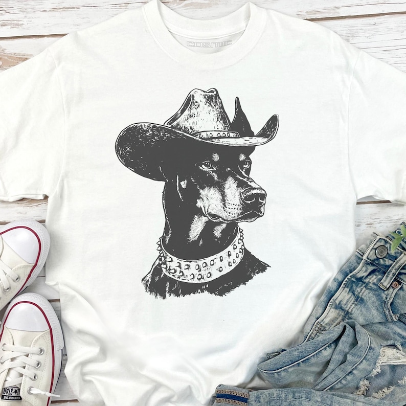 Pu&ograve; includere: T-shirt bianco con un'illustrazione in bianco e nero di un Dobermann Pinscher con cappello da cowboy e collare borchiato. Il ritratto del cane &egrave; centrato sulla maglietta. La maglietta &egrave; abbinata a jeans blu e scarpe da ginnastica bianche.