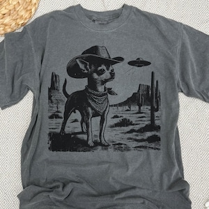 May include: Dark gray t-shirt featuring a black and white graphic of a Chihuahua wearing a cowboy hat and bandana, set against a desert landscape with cacti and a UFO. The shirt is made of a soft material.