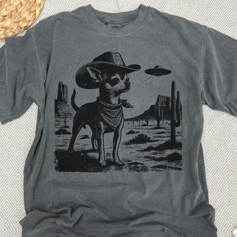 May include: Dark gray t-shirt featuring a black and white graphic of a Chihuahua wearing a cowboy hat and bandana, set against a desert landscape with cacti and a UFO. The shirt is made of a soft material.