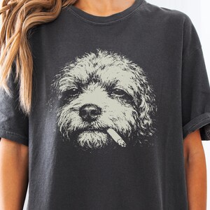 以下が含まれることがあります： チャコールグレーの半袖Tシャツ。口にタバコをくわえた巻き毛の犬の大きな明るい色のグラフィックプリントが施されています。クルーネックでゆったりとしたフィット感があり、コントラストの効いた詳細なイラストが特徴です。