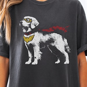 May include: Dark gray t-shirt featuring a white dog illustration with a superhero theme. The dog wears a yellow mask and a red cape. A yellow emblem is on the dog's chest. The shirt is a casual wear item.