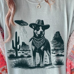 Peut inclure: T-shirt gris clair avec un dessin en noir et blanc d'un chien portant un chapeau de cowboy et un bandana, avec un OVNI au-dessus. Le motif comprend des éléments de paysage désertique tels que des cactus et des montagnes.