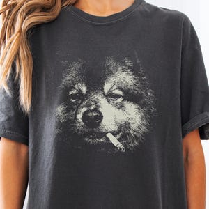 Może przedstawiać: Ciemnoszary t-shirt z krótkim rękawem z dużym, kontrastowym nadrukiem graficznym przedstawiającym puszystego psa z papierosem w pysku. Wzór utrzymany jest w odcieniach złamanej bieli i czerni, umieszczony centralnie na klatce piersiowej.