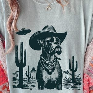 May include: A light gray t-shirt featuring a Boxer dog wearing a cowboy hat and bandana, with a UFO and desert landscape graphic. The design is in black and white, with cacti and mountains.