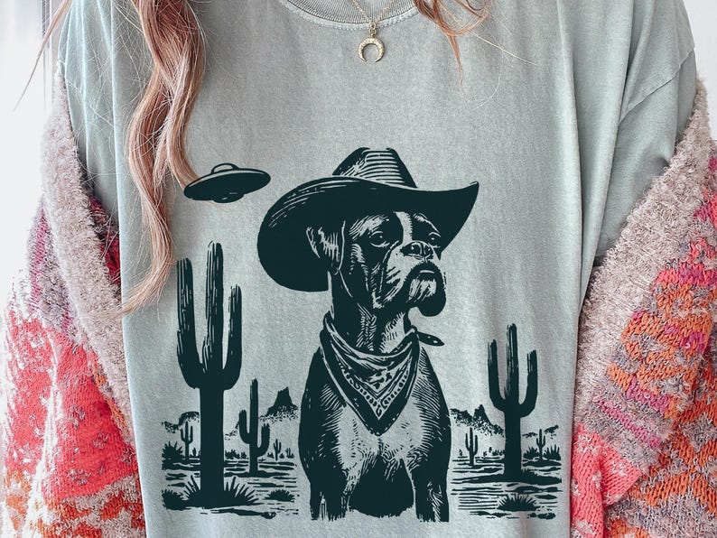 Puede incluir: Camiseta gris claro con un perro Boxer con sombrero de vaquero y pa&ntilde;uelo, con un OVNI y un paisaje des&eacute;rtico. El dise&ntilde;o es en blanco y negro, con cactus y monta&ntilde;as.