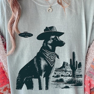 May include: A light blue t-shirt featuring a black and white illustration of a dog wearing a cowboy hat and bandana, with a UFO above and desert landscape in the background. The design is printed on a soft, comfortable tee.