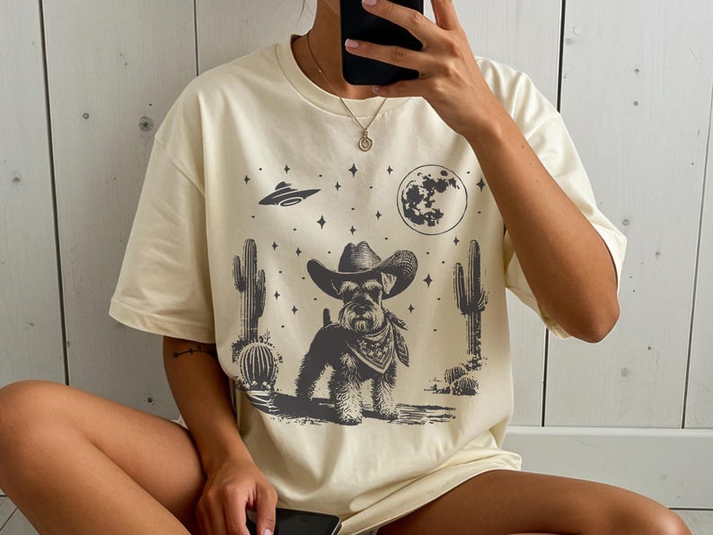 May include: Cream-colored t-shirt featuring a Schnauzer dog wearing a cowboy hat and bandana, set against a desert landscape with cacti, a full moon, stars, and a UFO. The graphic is in shades of gray.