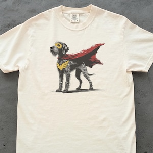 May include: Cream-colored t-shirt featuring a superhero dog graphic. The dog is depicted with a yellow mask, a red cape, and a gold emblem. The shirt is made of a soft, comfortable material, perfect for casual wear.