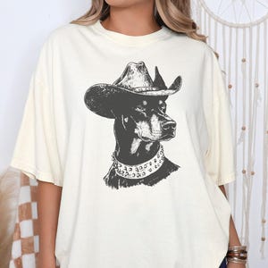 Pu&ograve; includere: T-shirt color crema con un'illustrazione in bianco e nero di un Dobermann Pinscher con cappello da cowboy e collare borchiato. La grafica &egrave; centrata sulla maglietta, offrendo un design unico ed elegante.