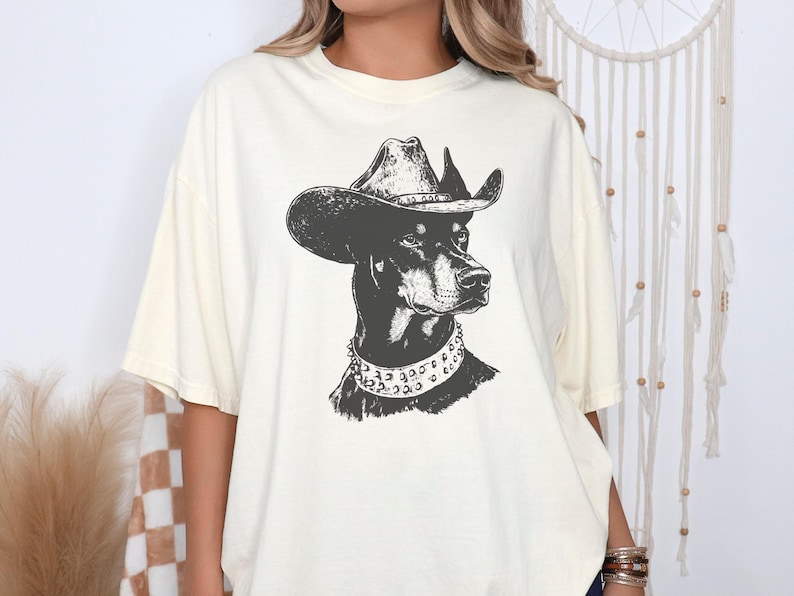 Pu&ograve; includere: T-shirt color crema con un'illustrazione in bianco e nero di un Dobermann Pinscher con cappello da cowboy e collare borchiato. La grafica &egrave; centrata sulla maglietta, offrendo un design unico ed elegante.