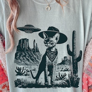 May include: A light blue t-shirt featuring a black and white graphic of a cat wearing a cowboy hat and bandana, with a desert landscape and a UFO. The cat is the central focus, with a detailed illustration style.