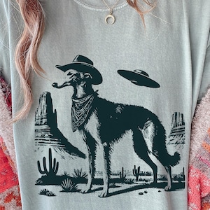Pode incluir: Camiseta azul clara com uma ilustração em preto e branco de um cão usando um chapéu de cowboy, bandana e fumando um cachimbo. O design inclui um OVNI e elementos da paisagem desértica.