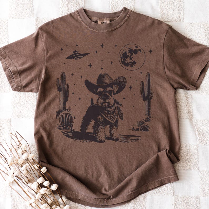May include: Brown t-shirt featuring a graphic of a dog wearing a cowboy hat and bandana, set against a desert landscape with cacti, a full moon, and a UFO. The design is in a vintage style.