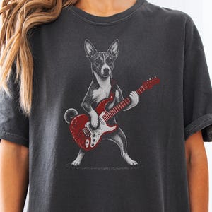 May include: A charcoal gray short-sleeve t-shirt featuring a graphic illustration of a dog standing on its hind legs and playing a red electric guitar. The dog is depicted in a black and white sketch style, contrasting with the vibrant red guitar.