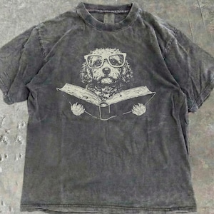 Può includere: Una t-shirt a maniche corte color grigio antracite con finitura effetto slavato. Al centro della maglia è presente un'illustrazione grafica di un cane dal pelo riccio che indossa occhiali dalla montatura spessa e tiene un libro aperto.