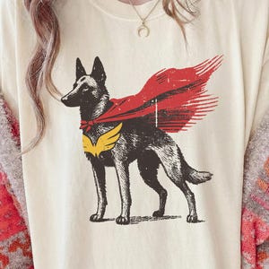 Puede incluir: Camiseta color crema con un gráfico de un perro con capa y emblema de superhéroe. El perro es negro y marrón, con una capa roja y un emblema amarillo. El diseño tiene un aspecto vintage y desgastado.