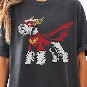 May include: Dark gray t-shirt featuring a cartoon Schnauzer dressed as a superhero. The dog wears a red cape, mask, and shirt with a gold emblem. The design has a vintage, distressed look.