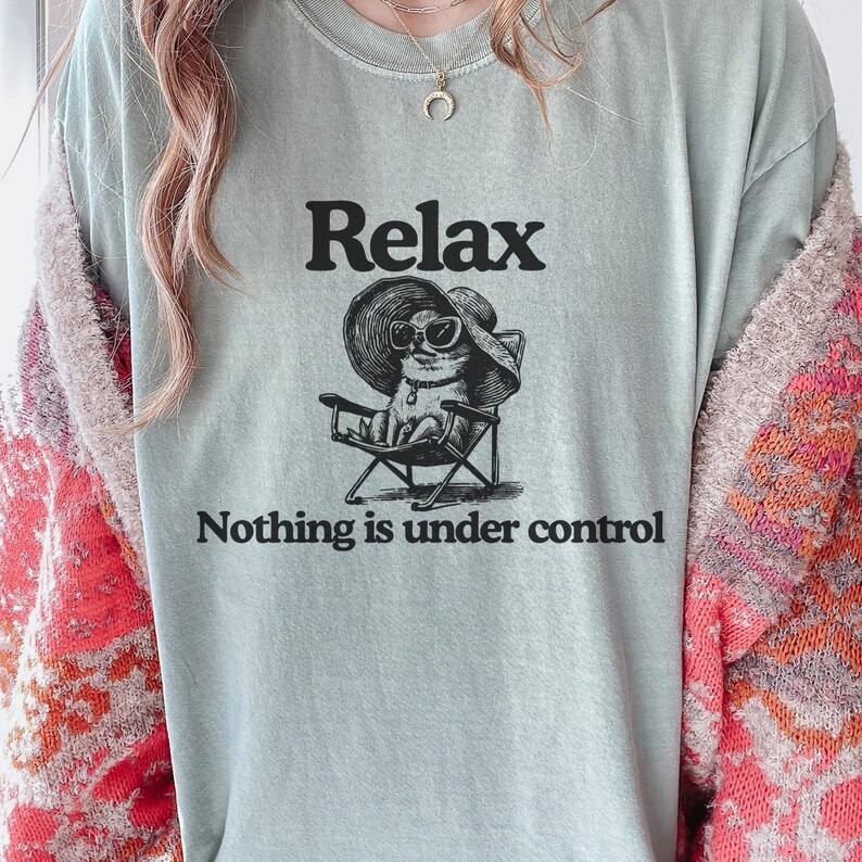 May include: A light green t-shirt with the word "Relax" above a graphic of a cat wearing sunglasses and a sun hat, seated in a folding chair. The text "Nothing is under control" is below the graphic.