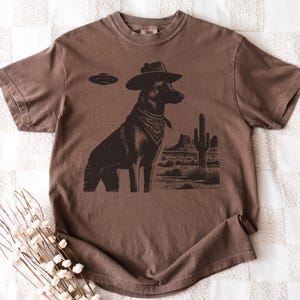 May include: Brown t-shirt featuring a graphic of a dog wearing a cowboy hat and bandana, with a UFO above and a desert landscape in the background. The shirt is a solid brown color.