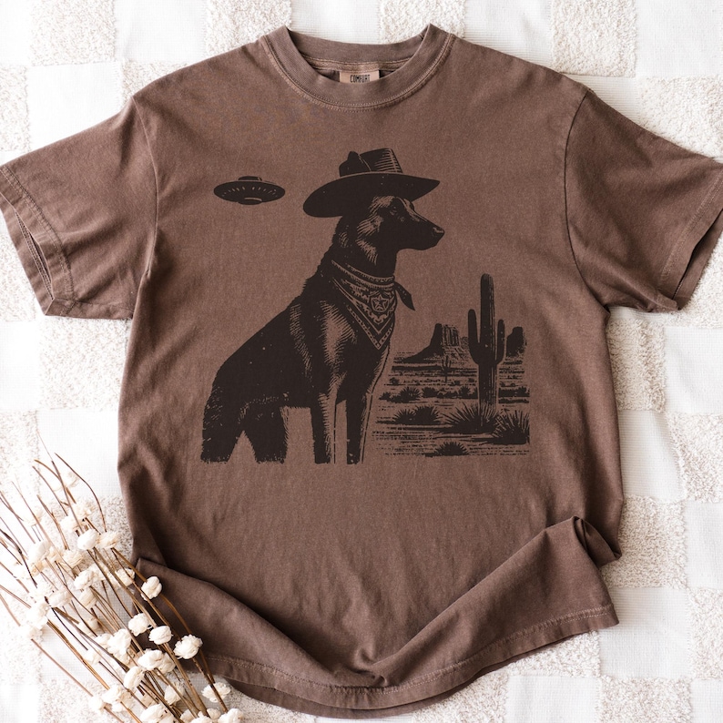 May include: Brown t-shirt featuring a graphic of a dog wearing a cowboy hat and bandana, with a UFO above and a desert landscape in the background. The shirt is a solid brown color.