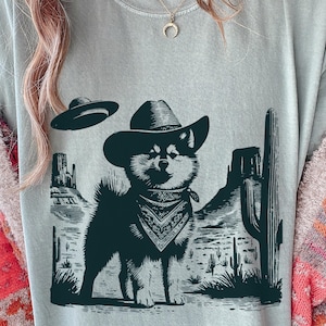 Pode incluir: Camiseta cinza claro com uma ilustração em preto e branco de um cachorro usando um chapéu de cowboy e bandana, em uma paisagem desértica. Um OVNI paira sobre o cachorro. O design inclui cactos e montanhas desérticas.