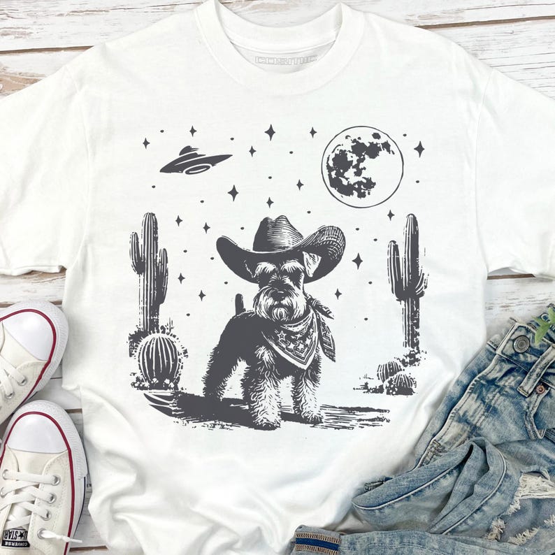 May include: White t-shirt featuring a black and white graphic of a Schnauzer dog wearing a cowboy hat and bandana, set against a desert landscape with cacti, a full moon, and a UFO. The shirt is paired with denim jeans and white sneakers.
