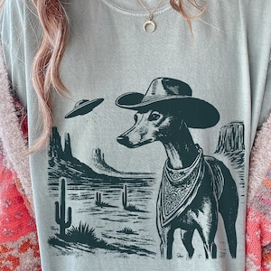 May include: A pale blue t-shirt with a monochrome graphic. It depicts a dog in a cowboy hat and bandana, set in a desert scene with a UFO. The design includes cacti and rock formations.