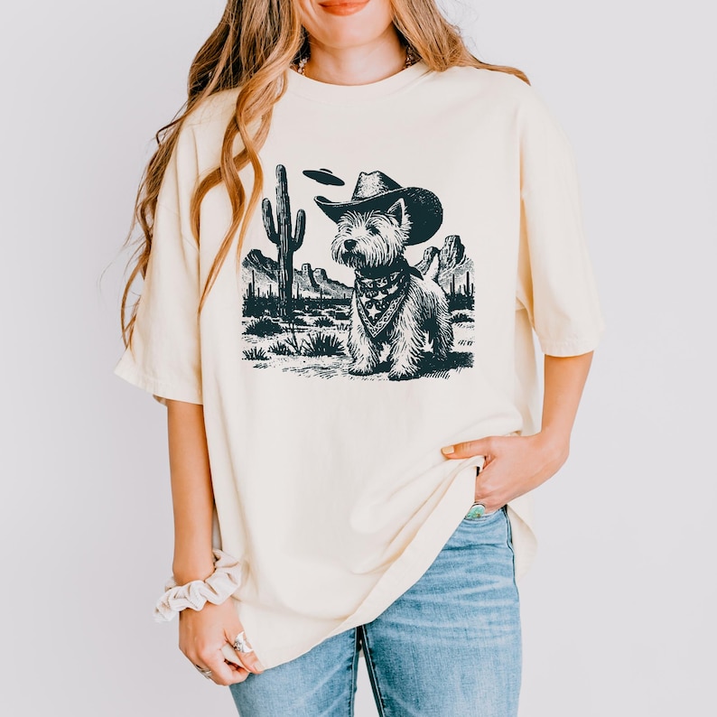May include: Cream-colored t-shirt featuring a black and white illustration of a dog wearing a cowboy hat and bandana, set against a desert landscape with cacti and mountains. A UFO hovers above the dog.