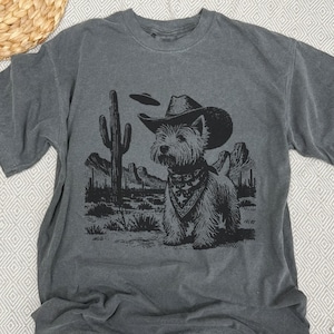 May include: Gray t-shirt featuring a black graphic of a dog wearing a cowboy hat and bandana in a desert landscape with a cactus and UFO. The dog is the central focus of the design.