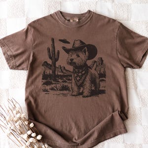 May include: Brown t-shirt featuring a graphic of a dog wearing a cowboy hat and bandana, set against a desert landscape with a cactus and mountains. The dog is the focal point of the design.