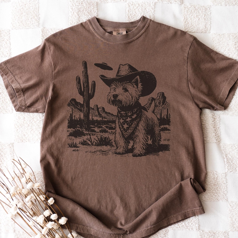 May include: Brown t-shirt featuring a graphic of a dog wearing a cowboy hat and bandana, set against a desert landscape with a cactus and mountains. The dog is the focal point of the design.