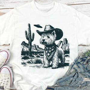 May include: White t-shirt featuring a West Highland White Terrier wearing a cowboy hat and bandana, set against a desert landscape with a cactus and UFO. The graphic is in shades of green and black.