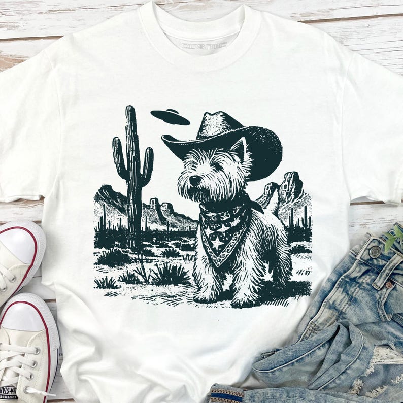 May include: White t-shirt featuring a West Highland White Terrier wearing a cowboy hat and bandana, set against a desert landscape with a cactus and UFO. The graphic is in shades of green and black.