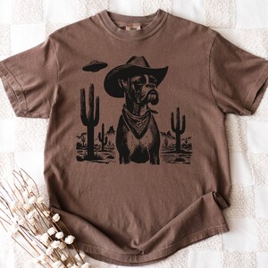 Puede incluir: Camiseta marr&oacute;n con un gr&aacute;fico negro de un perro con sombrero de vaquero y pa&ntilde;uelo, con un OVNI y un paisaje des&eacute;rtico. Cuello redondo y mangas cortas.
