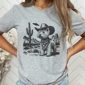 May include: A light gray t-shirt featuring a black and white graphic of a dog wearing a cowboy hat and bandana, set against a desert landscape with cacti and mountains. The dog is the central focus of the design.