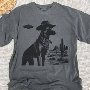May include: Dark gray t-shirt featuring a black graphic of a dog wearing a cowboy hat and bandana, with a UFO above and a desert landscape. The shirt is made of a soft, comfortable material.