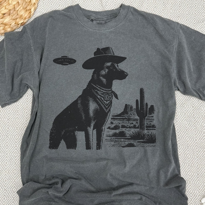 May include: Dark gray t-shirt featuring a black graphic of a dog wearing a cowboy hat and bandana, with a UFO above and a desert landscape. The shirt is made of a soft, comfortable material.