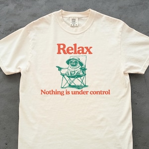 Könnte beinhalten: Cremefarbenes T-Shirt mit dem Wort "Relax" in Orange über einer grünen Illustration eines Mops mit Hut, der in einem Stuhl sitzt. Der Text "Nothing is under control" steht darunter.