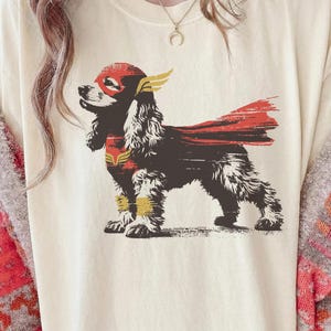 Op de afbeelding: Crèmekleurig T-shirt met een afbeelding van een zwart-bruine Cocker Spaniel in een superheldenpak. De hond draagt een rood masker en cape met gele accenten. Het ontwerp is een speelse interpretatie van het superheldenthema.