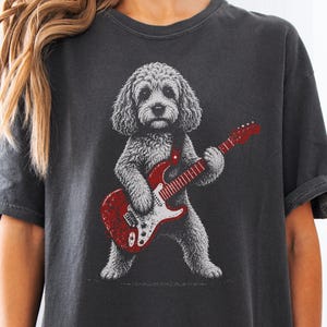 Peut inclure: Un t-shirt gris anthracite à manches courtes avec une impression graphique représentant un chien au pelage touffu debout sur ses pattes arrière, jouant d'une guitare électrique rouge vif. Le chien a un aspect texturé, façon croquis.