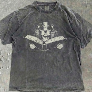 May include: A charcoal grey, acid-washed short-sleeve t-shirt featuring a graphic print of a dog wearing white-rimmed glasses while holding an open book. The dog is centered on the chest, and the shirt has a crew neckline and a relaxed fit.