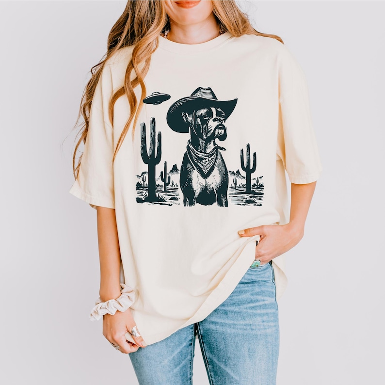 Puede incluir: Camiseta color crema con un gr&aacute;fico en blanco y negro de un perro Boxer con sombrero de vaquero y pa&ntilde;uelo, con un OVNI y cactus en el fondo. La camiseta tiene un corte holgado y mangas cortas.