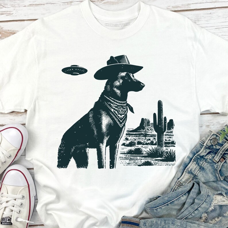 May include: White t-shirt featuring a dark green graphic of a dog wearing a cowboy hat and bandana, with a UFO above. A desert landscape with a cactus and mountains is in the background.