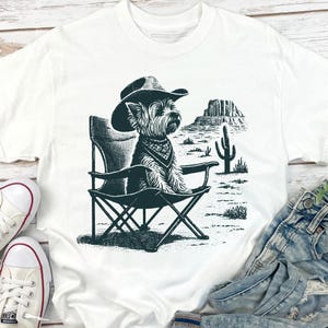 Pu&ograve; includere: T-shirt bianco con un'illustrazione in bianco e nero di un cane con cappello da cowboy e bandana, seduto su una sedia pieghevole. Lo sfondo raffigura un paesaggio desertico con un cactus e formazioni rocciose. Abbigliamento casual.